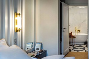 Quarto de hotel com uma cama, mesa de cabeceira com revistas, porta que dá para um banheiro com banheira e chuveiro.