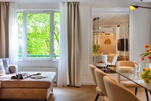 A modern hotel room with a bed, a dining area with chairs and a table, a TV, and a view of greenery outside.