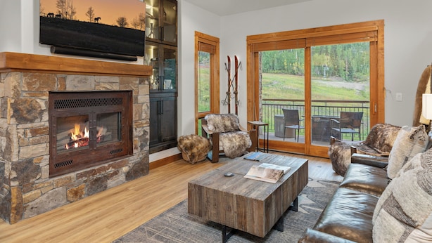 A cozy living room with a stone fireplace, leather sofa, and a large window overlooking a grassy area. - Terraces A+ TRUE Ski-In Ski-Out 2 Bedroom, 2 Bathrooms - Sleeps 4 (Mountain Village)