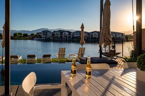 An infinity pool with a view of houses across the water, a table with two bottles of Corona beer, and lounge chairs. - The Lake House - Terra Lago (Indio)