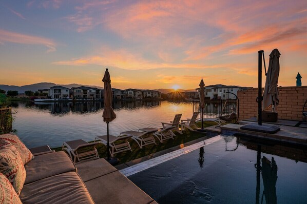 A poolside area with lounge chairs and umbrellas, a clear view of the sunset over the water, and a row of buildings in the background. - The Lake House - Terra Lago (Indio)