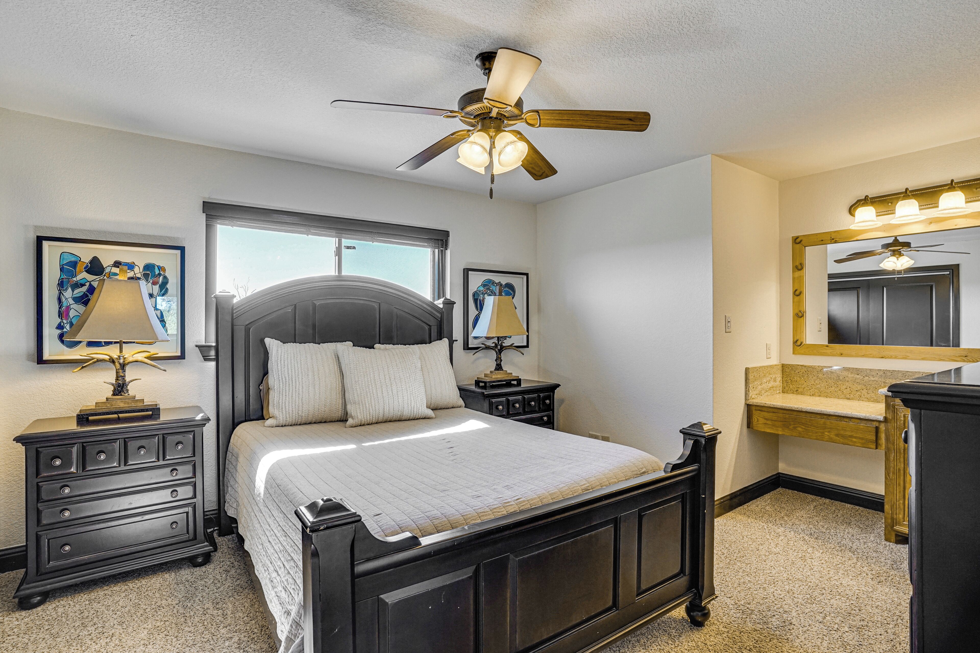 A bedroom with a bed, a ceiling fan, bedside tables with lamps, a window, and a bathroom with a vanity and mirror.