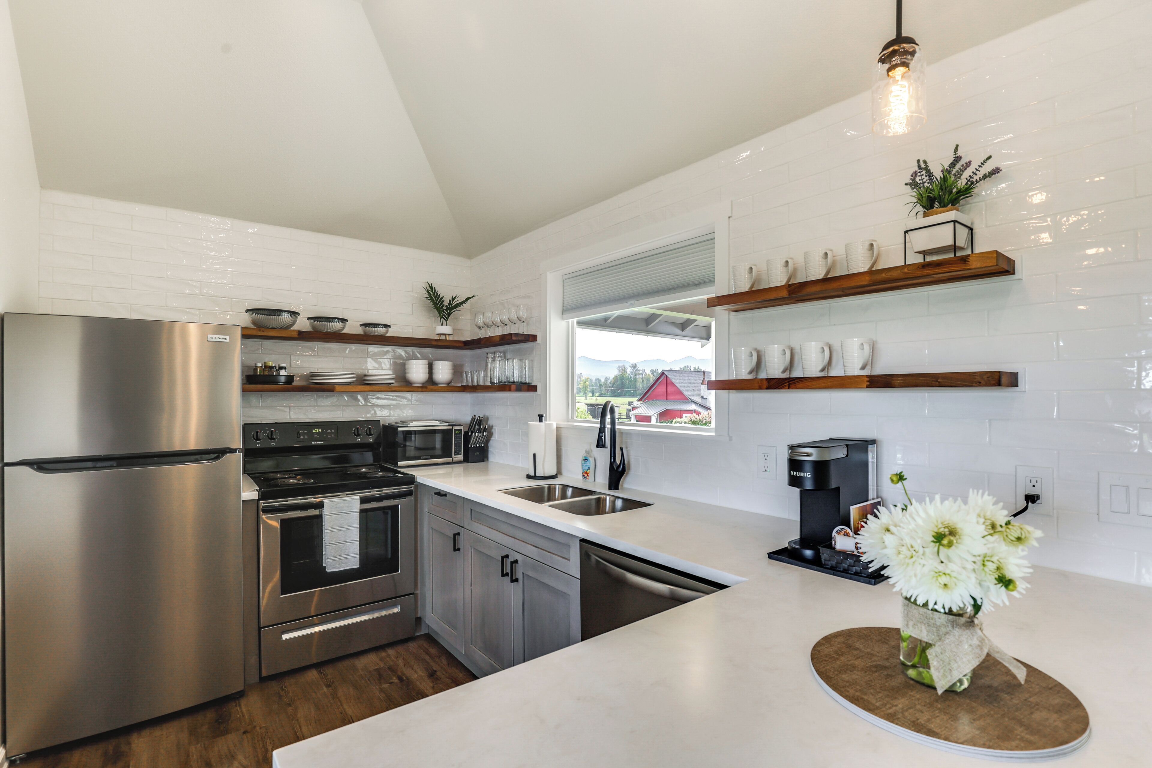 Una cocina moderna con electrodomésticos de acero inoxidable, estantes de madera y vistas a una casa con techo rojo.