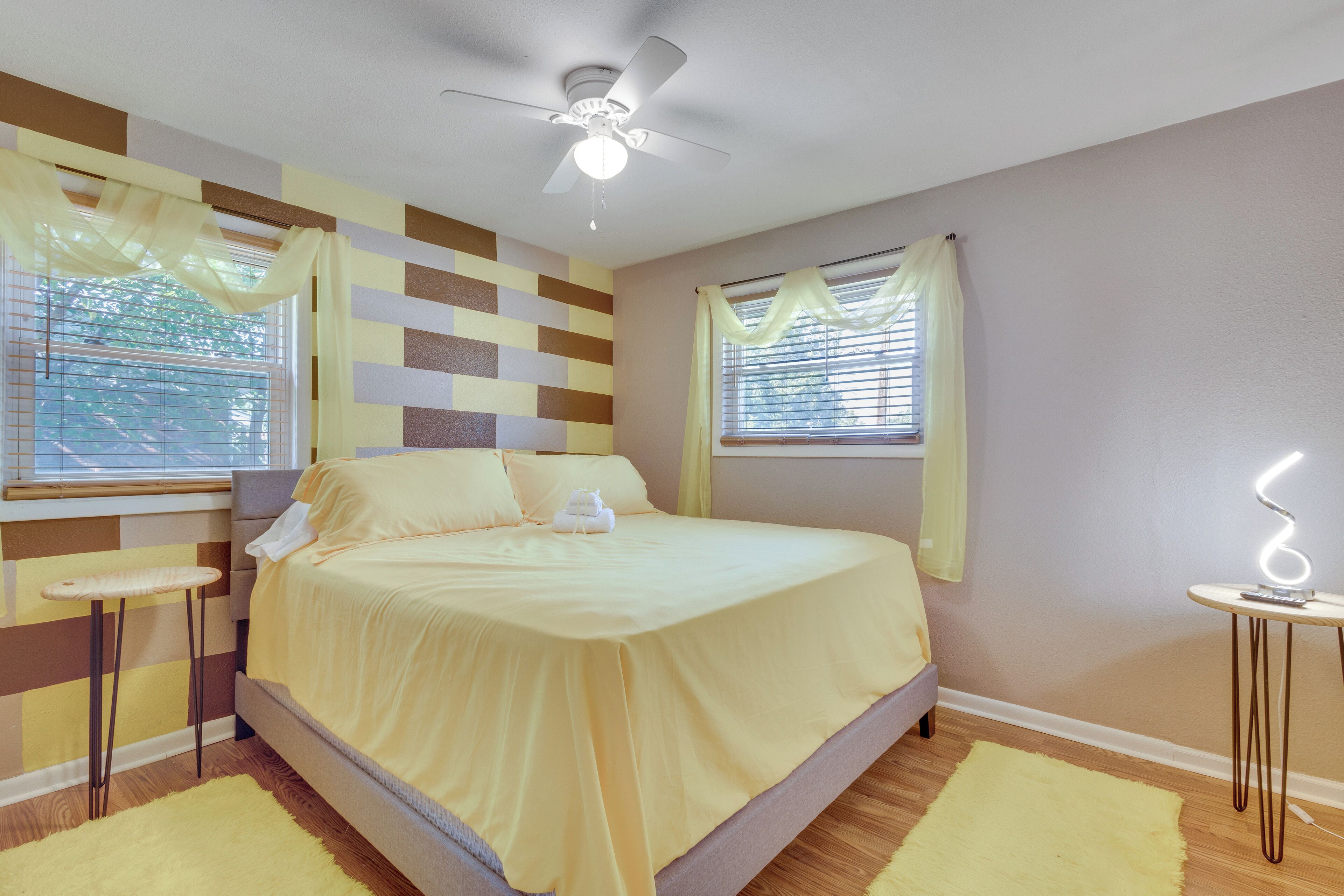 A bedroom with a bed, a ceiling fan, a bedside table, a window with curtains, and a small table with a light.