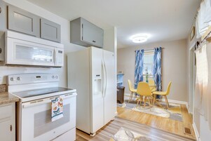 Cocina compacta con microondas, horno y refrigerador. Cuenta con una zona de comedor con mesa y sillas, y una ventana con cortinas.