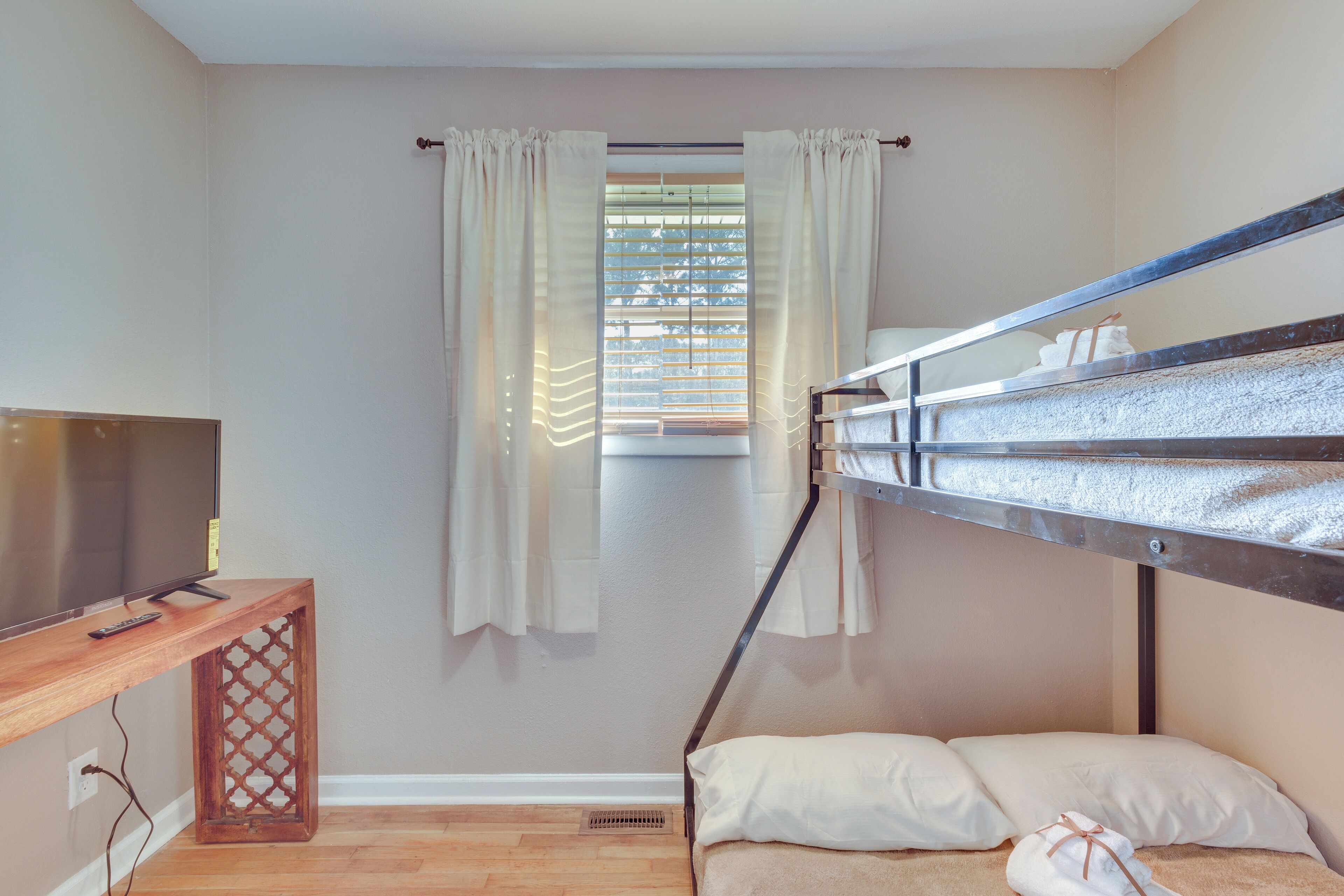 Una cama individual sobre otra, con un televisor sobre una mesa consola de madera, una ventana con cortinas y un radiador.