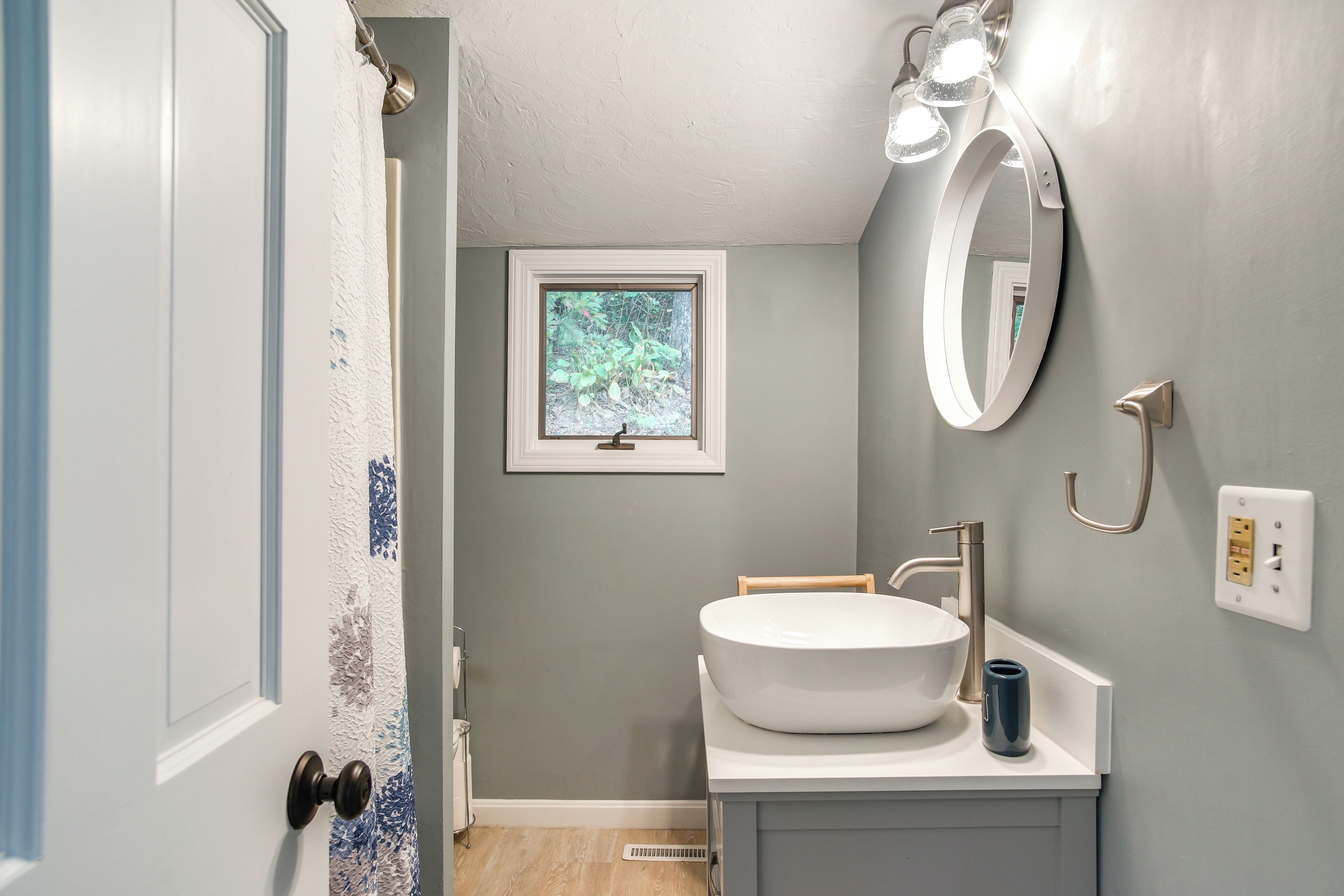 A bathroom with a white sink, a mirror, and a window with a view of greenery.