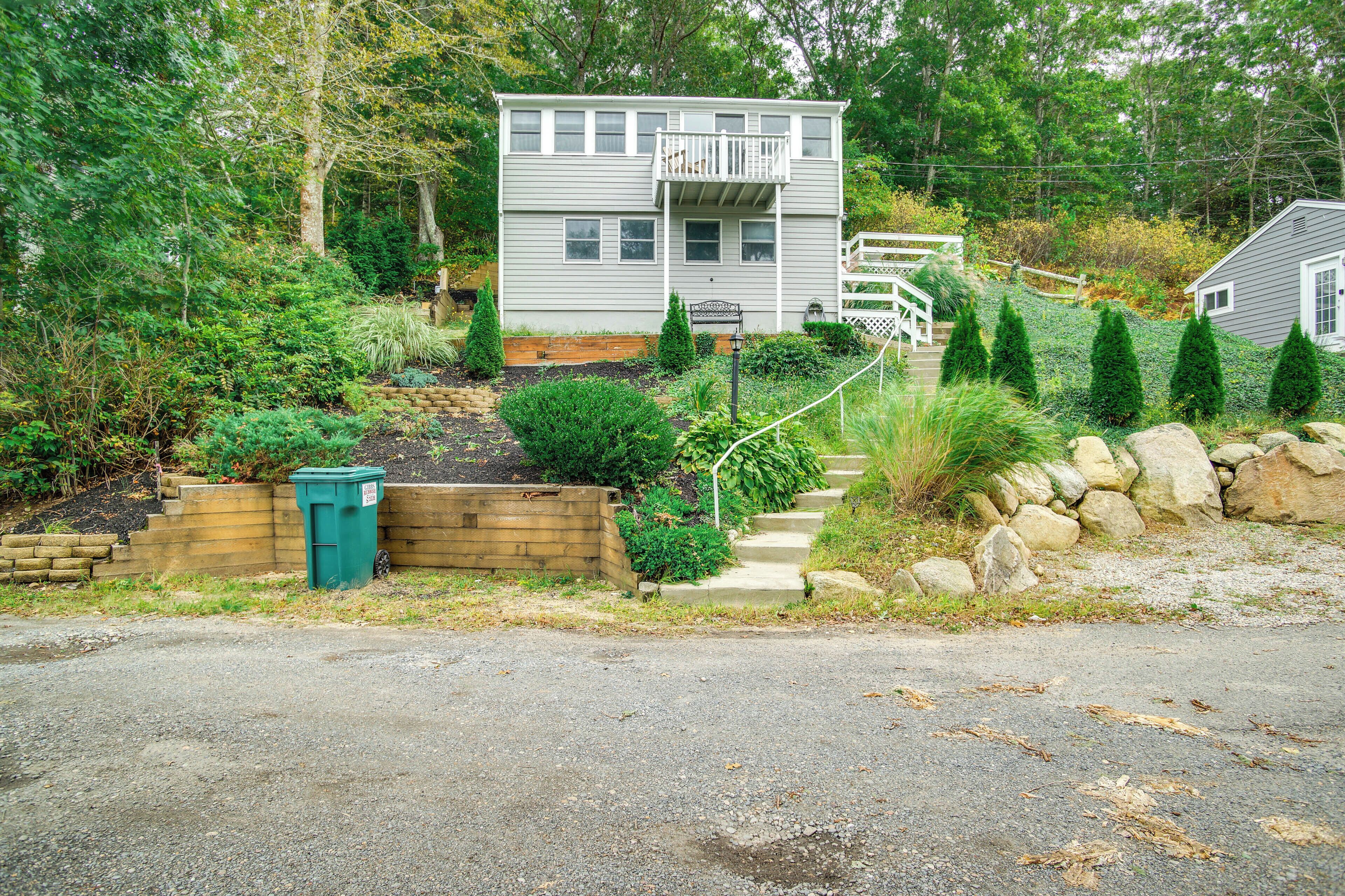 A two-story house with a balcony, surrounded by a garden with various plants and a stone wall.