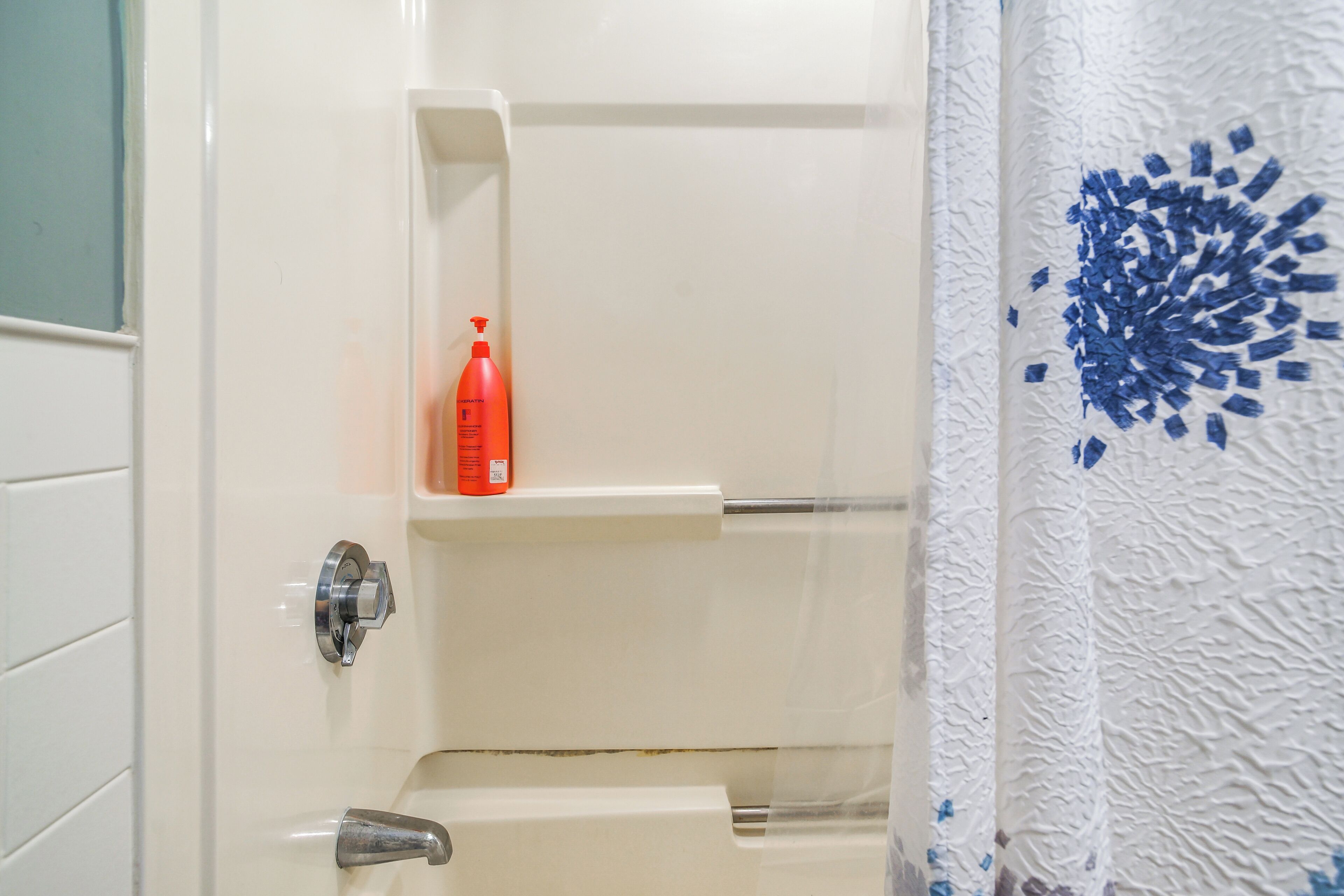 Ein Badezimmer mit einer weißen Badewanne, einer roten Flasche auf einem Regal und einem Duschvorhang mit blauem Muster.