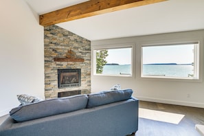 A living room with a stone fireplace, a gray sofa, and large windows overlooking a body of water. - Waterfront Roque Bluffs Hideaway w/ Private Beach (Machias)