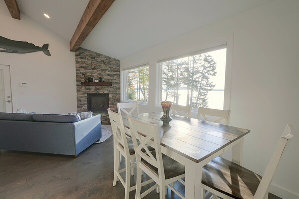 A dining area with a stone fireplace, a blue sofa, and a view of trees through large windows. - Waterfront Roque Bluffs Hideaway w/ Private Beach (Machias)