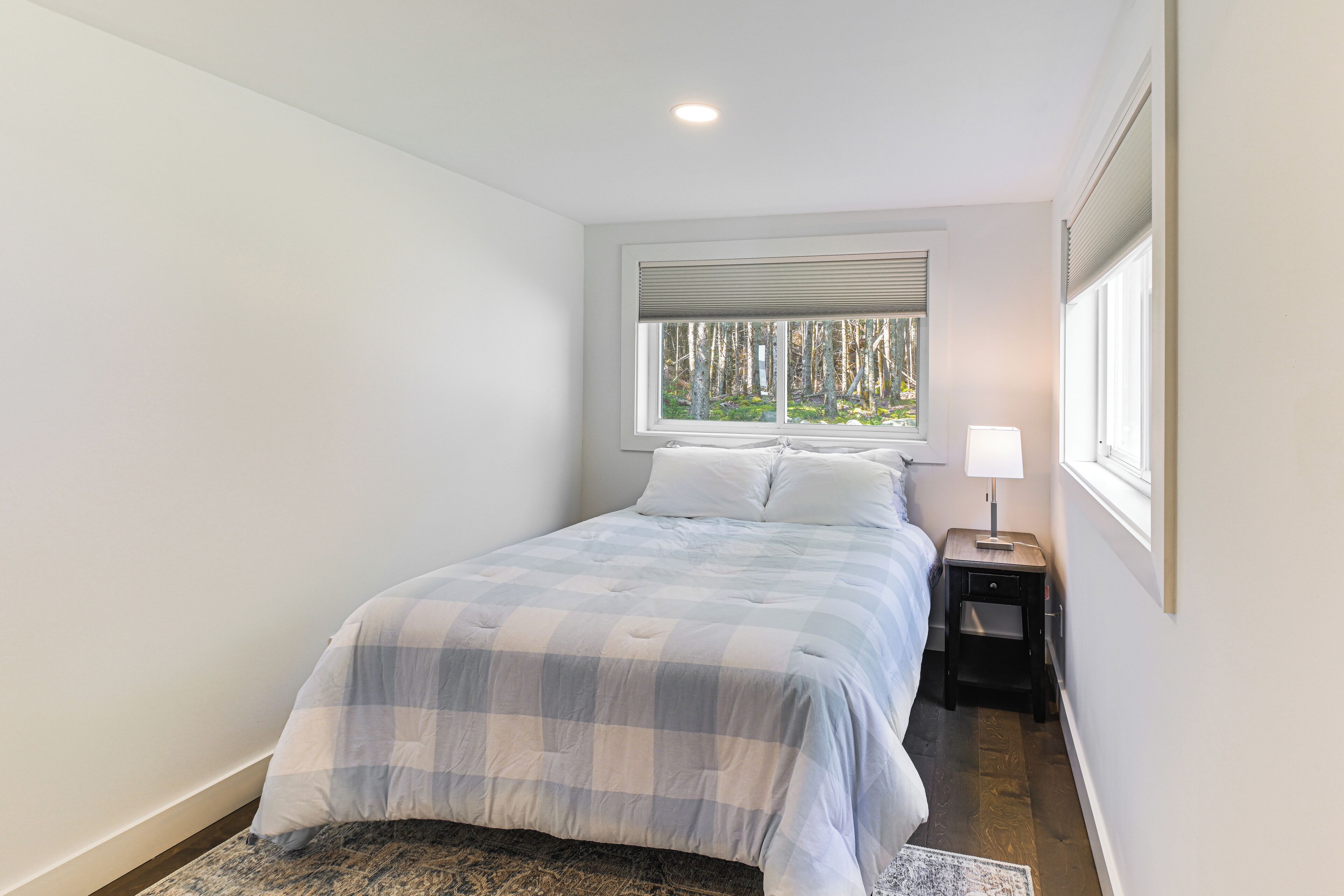 Une chambre à coucher comprenant un lit, une table de chevet, une lampe, une fenêtre avec des stores et une vue sur les arbres à l’extérieur.
