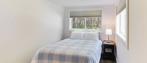 Une chambre à coucher comprenant un lit, une table de chevet, une lampe, une fenêtre avec des stores et une vue sur les arbres à l’extérieur.