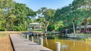 A wooden dock extending into a calm body of water, surrounded by lush greenery and a building with a green roof. - Waterfront Gulf Coast Home w/ Outdoor Oasis! (Gautier)