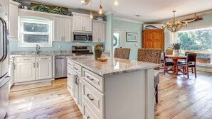 A spacious kitchen with white cabinetry, a central island, and a dining area with wooden chairs. - Waterfront Gulf Coast Home w/ Outdoor Oasis! (Gautier)