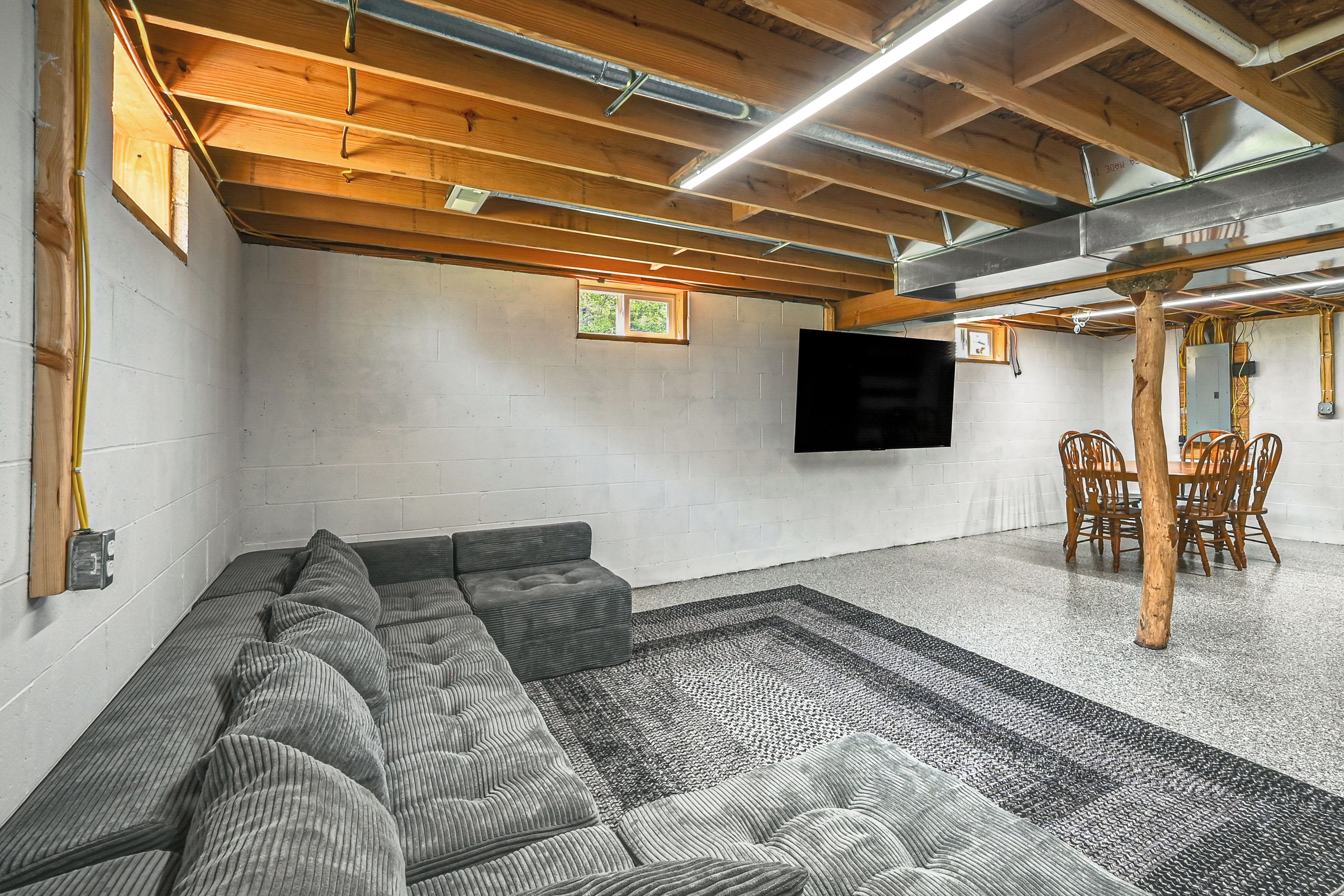 A room with a sectional sofa, a dining table with chairs, a flat-screen TV mounted on the wall, and exposed wooden beams on the ceiling.