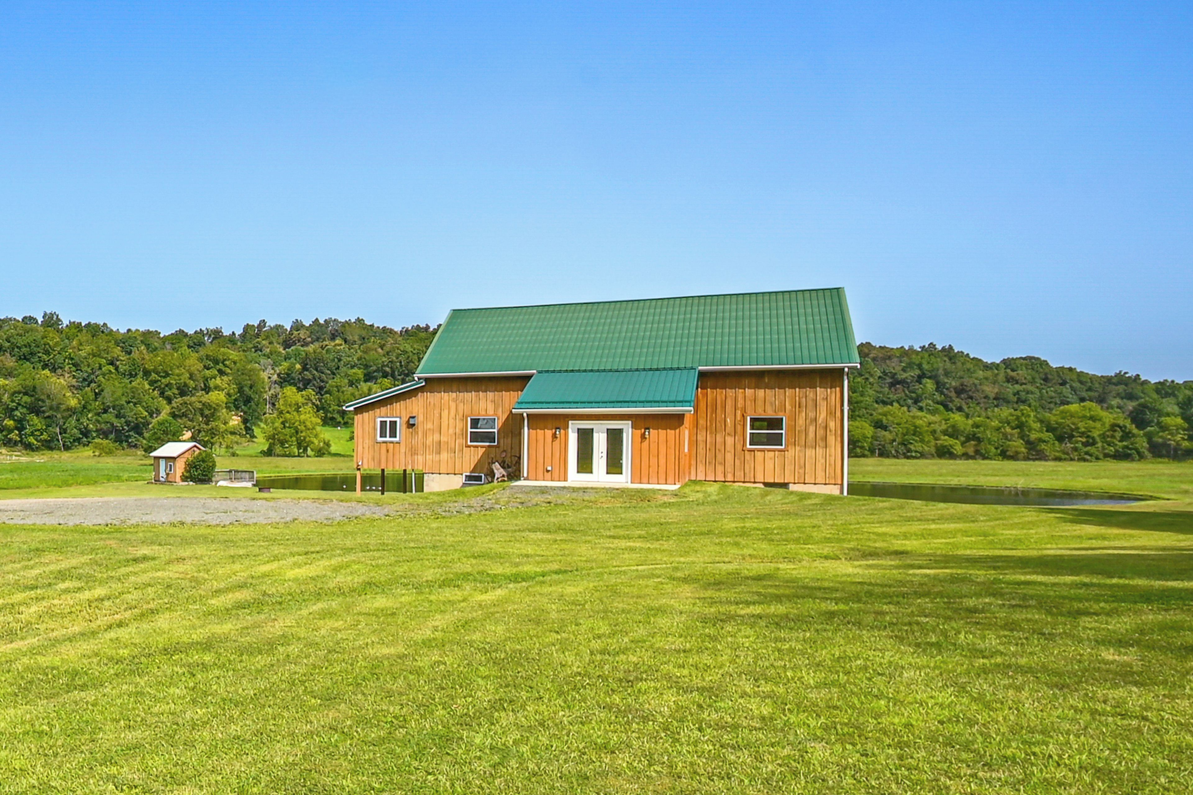 A two-story wooden house with a green roof, surrounded by a well-maintained lawn and a forested hill in the background.