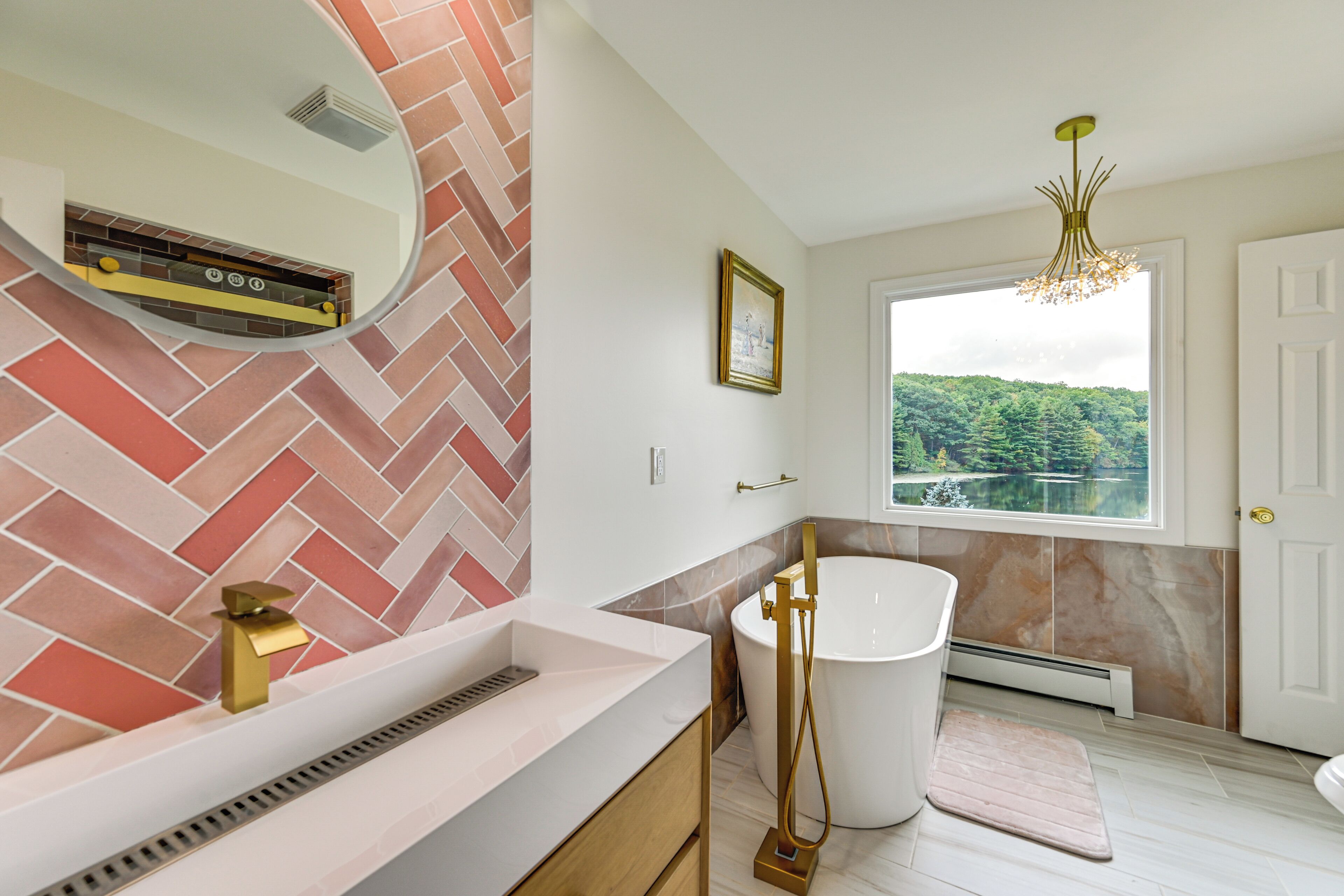 A modern bathroom with a herringbone tile backsplash, a freestanding bathtub, and a large window offering a view of a lake.