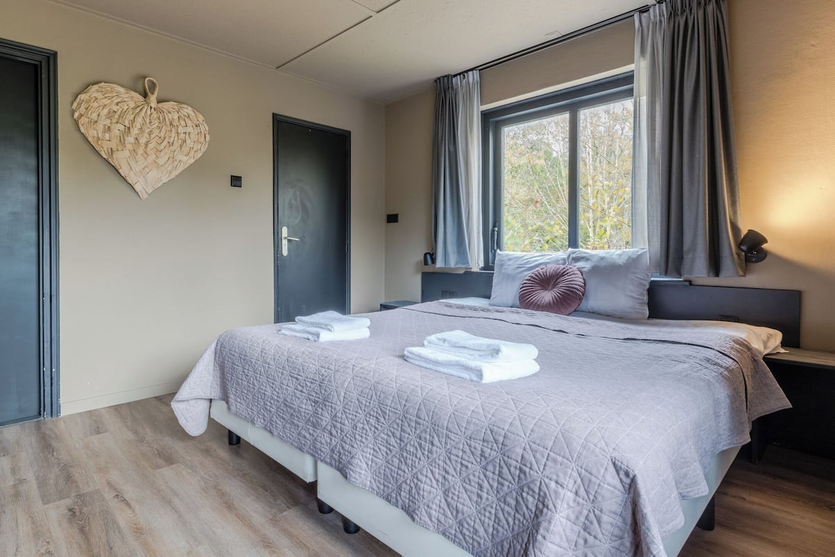 A modern bedroom with a large bed, a heart-shaped wall decoration, a window with curtains, and a nightstand with a lamp.