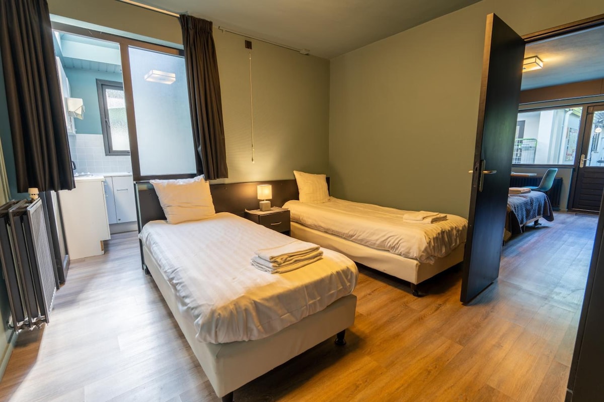 A hotel room with two beds, a small bedside table, a lamp, and a view of the street through an open door.