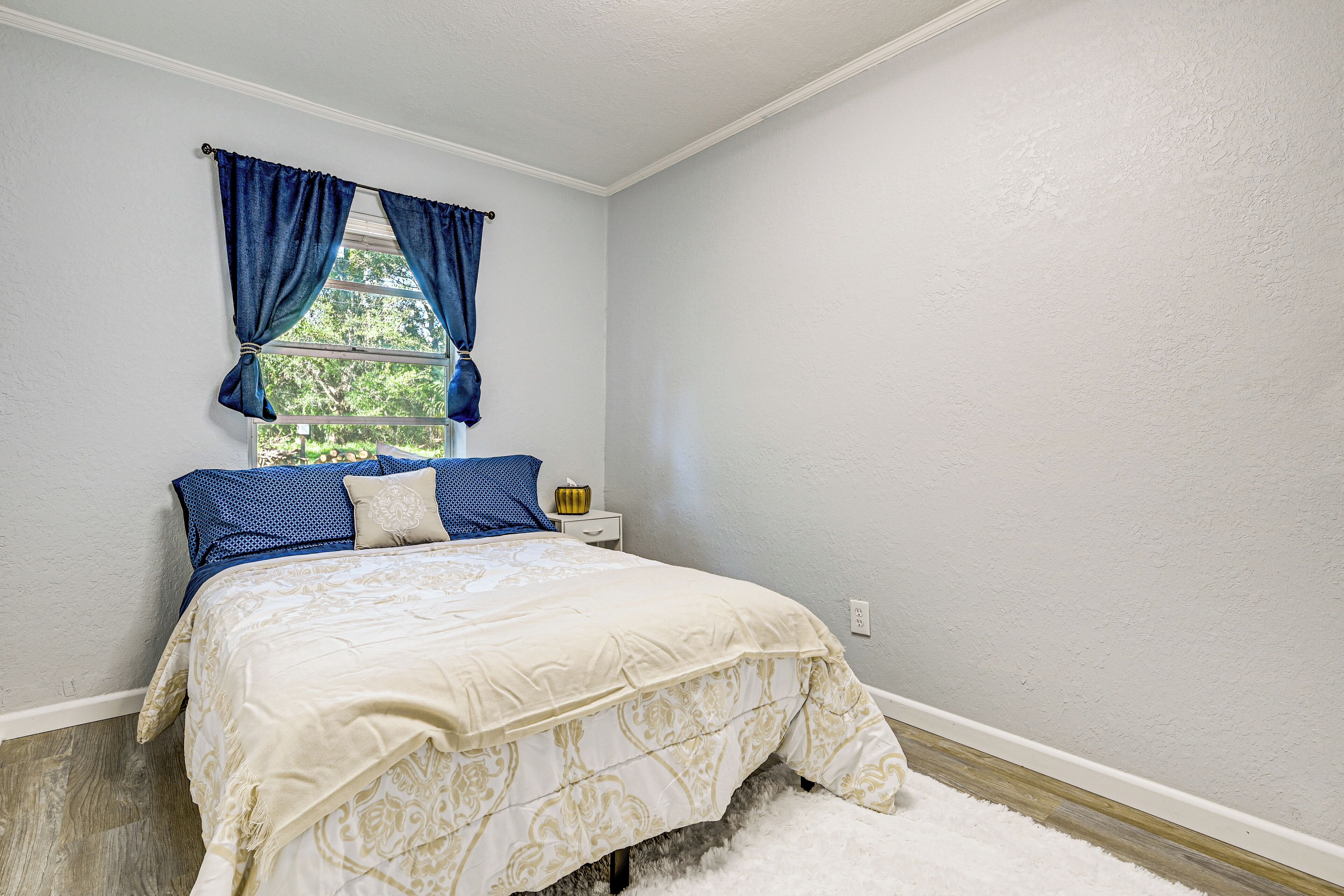 A bedroom with a bed, a window with blue curtains, a nightstand with a lamp, and a white wall.