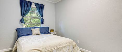 A bedroom with a bed, a window with blue curtains, a nightstand with a lamp, and a white wall.
