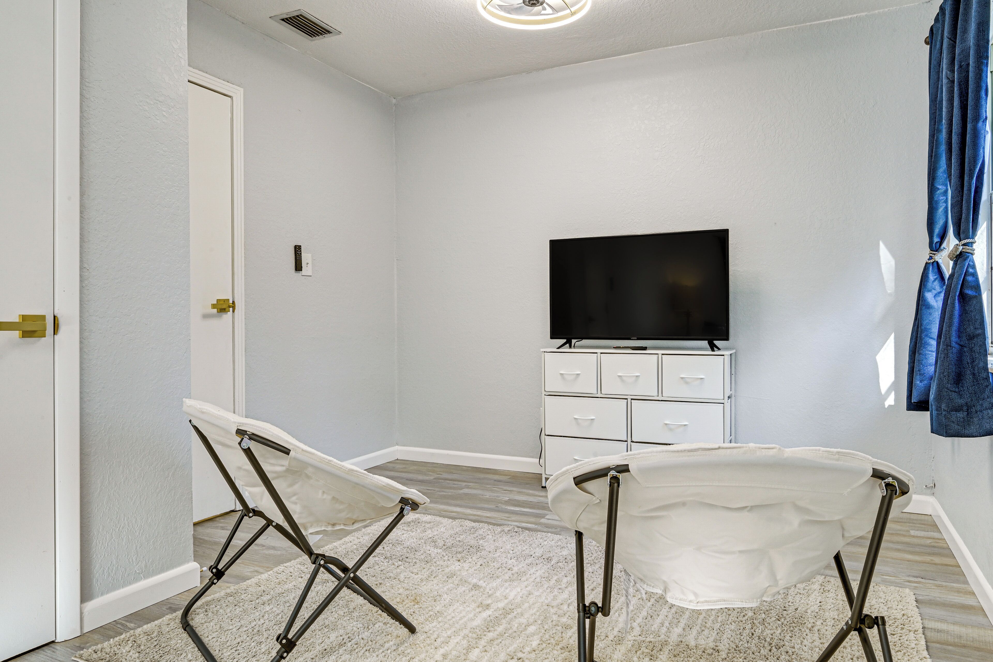 Una habitación con pared blanca, un televisor de pantalla plana sobre un tocador blanco, dos sillas plegables y una alfombra blanca.