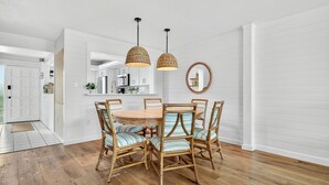 A dining area with a round wooden table, six wicker chairs with striped cushions, two woven pendant lights, and a round mirror on the wall. - Stunning ocean view condo in Sanibel, 2BR, 2BA, perfect for up to 6 guests. (Sanibel)