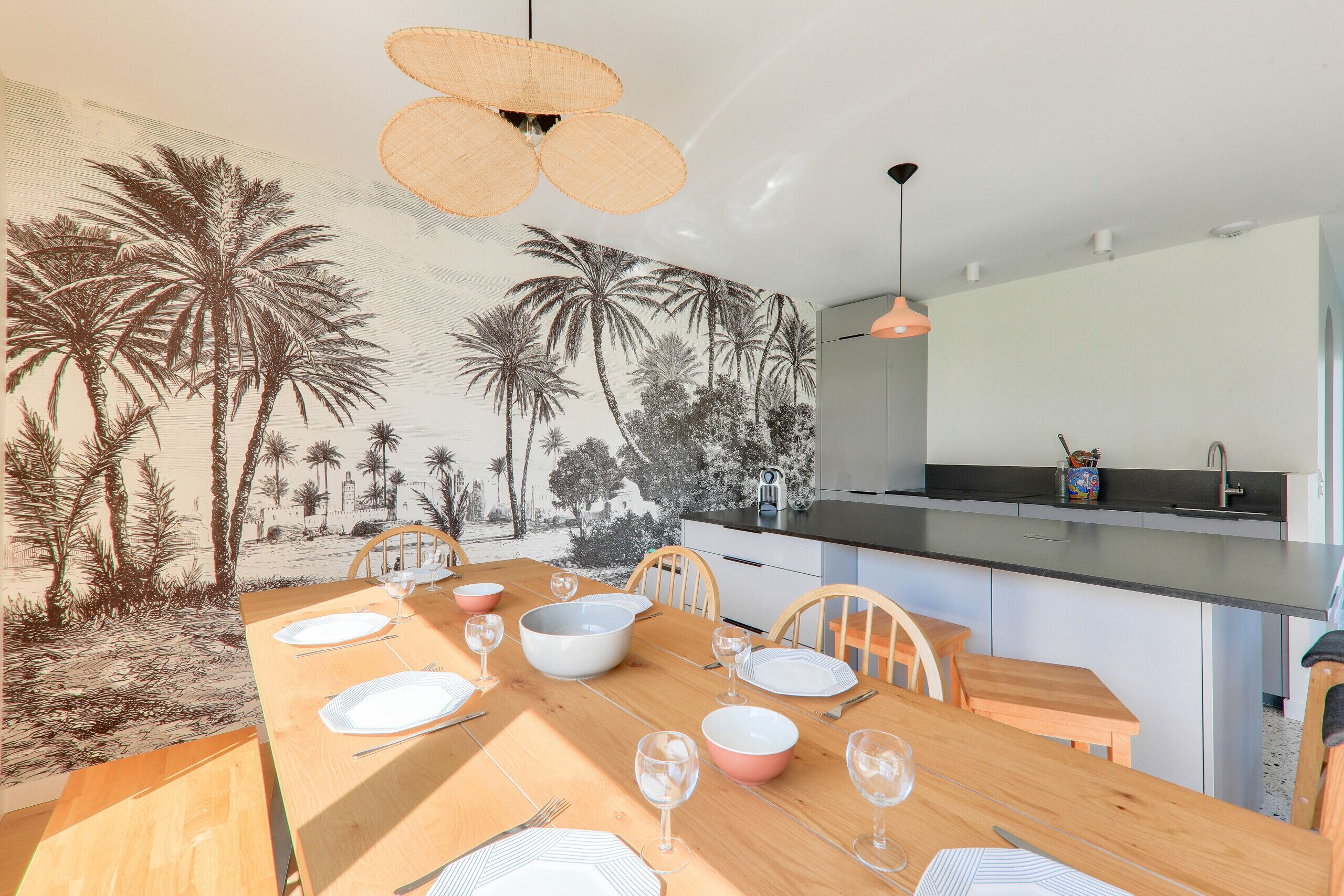 A dining area with a wooden table set for six, a mural of palm trees, and a modern kitchen in the background.