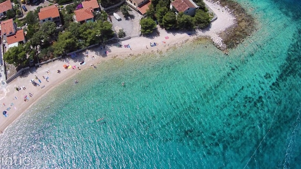 An aerial view of a coastal area with a beach, clear turquoise waters, and a cluster of buildings with red-tiled roofs. - Holiday Home Nada Rogoznica (Rogoznica)