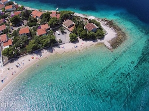 An aerial view of a coastal area with a beach, clear turquoise waters, and a cluster of buildings with red-tiled roofs. - Holiday Home Nada Rogoznica (Rogoznica)