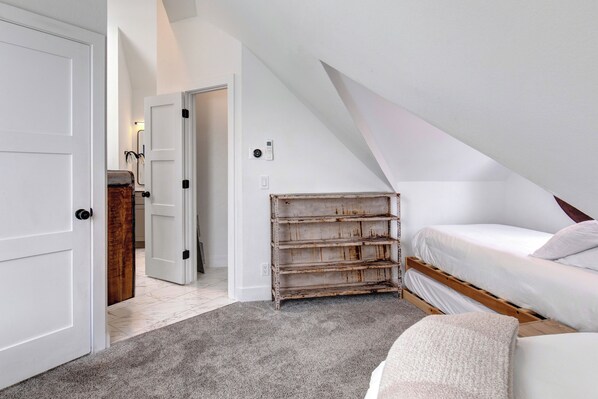 Un dormitorio con techo inclinado, una cama, un estante de madera y una puerta que da a otra habitación.