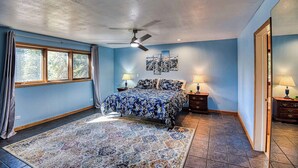 Une chambre à coucher comprenant un lit, un ventilateur de plafond, deux tables de chevet avec des lampes, une grande fenêtre avec des rideaux et un tapis.