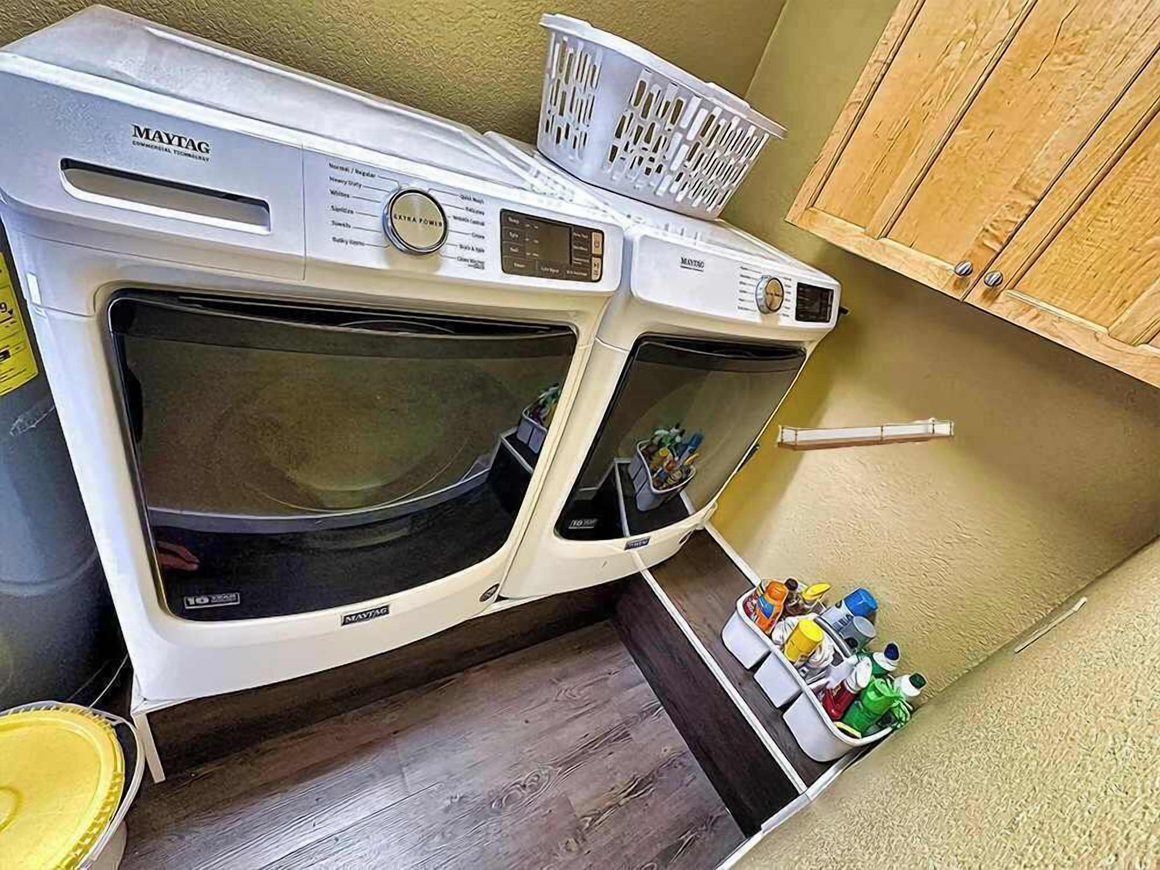 Une petite buanderie équipée de deux lave-linge Maytag à chargement frontal, un panier posé sur le lave-linge du dessus et une étagère contenant des produits de nettoyage.