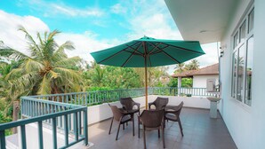 A balcony with wicker furniture and a green umbrella, overlooking a lush garden and palm trees.