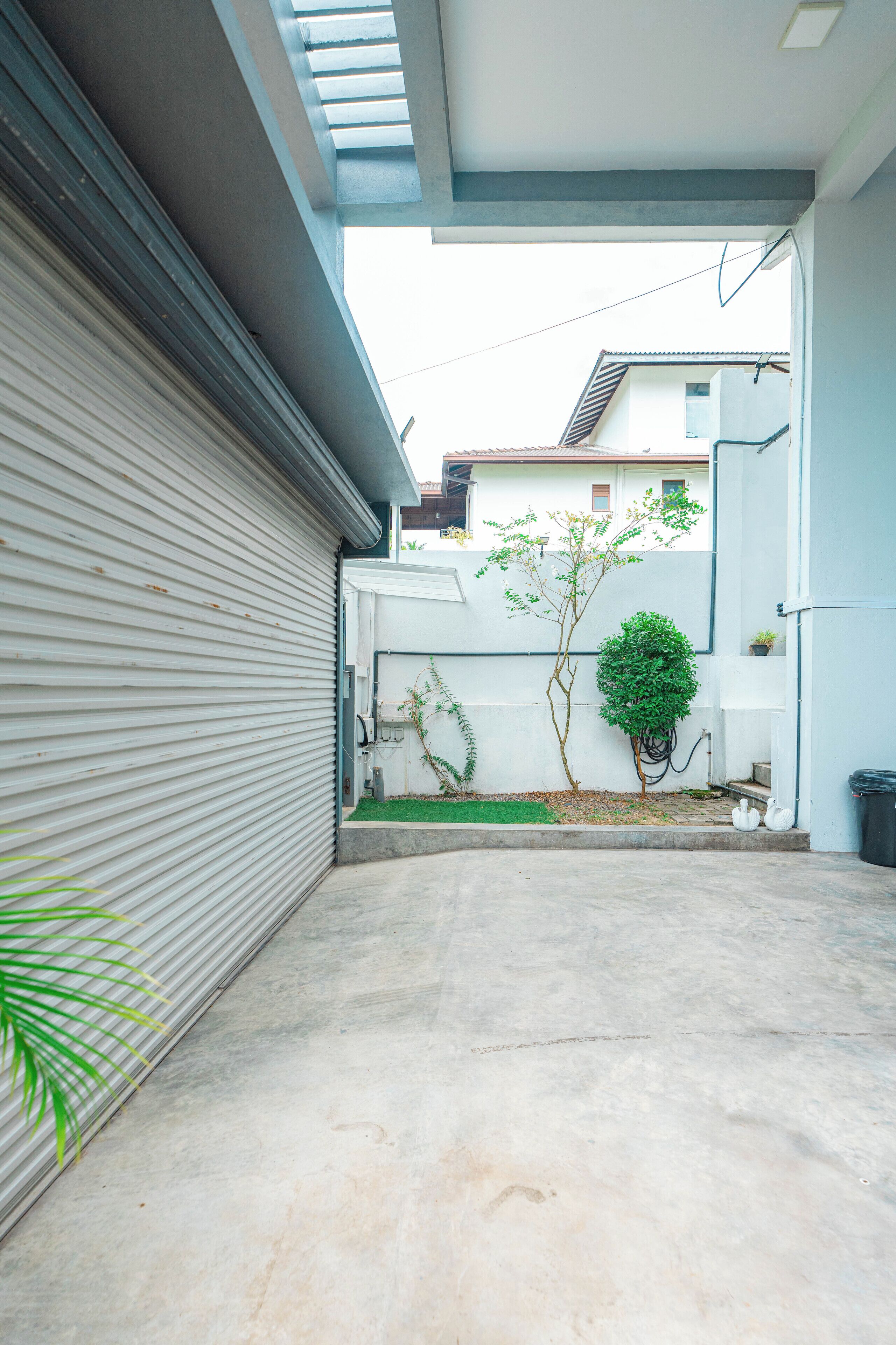 Une cour étroite et pavée, avec une porte de garage fermée, un petit arbre et un mur blanc.
