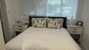 A neatly made bed with a white quilt, two nightstands with lamps, and a window with blinds. - Charming 1-bedroom apartment in delightful Biscayne Park (North Miami)