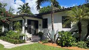 A single-story house with a white exterior, a black front door, and a small porch with a white bench. - Charming 1-bedroom apartment in delightful Biscayne Park (North Miami)
