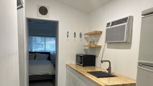 A compact kitchen with a microwave, sink, and built-in air conditioner. A bed is visible in the adjacent room, and there are decorative items on the wall. - Charming 1-bedroom apartment in delightful Biscayne Park (North Miami)