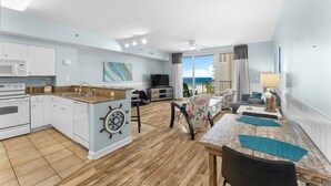 A modern kitchen with white cabinets, a microwave, and a coffee maker. There is a dining area with a wooden table and chairs, and a view of the ocean through large windows. - Waterfront condo w/ pool & beach access (Panama City Beach)