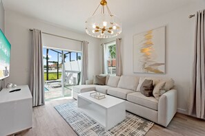 A modern living room with a white sofa, a coffee table, and a chandelier. There is a large window with curtains, a painting on the wall, and a TV on a stand. - Boutique Retreat, Azzure Resort, Near Orlando And Disney By Shine Villas 6 Bedroom Townhouse (Davenport)