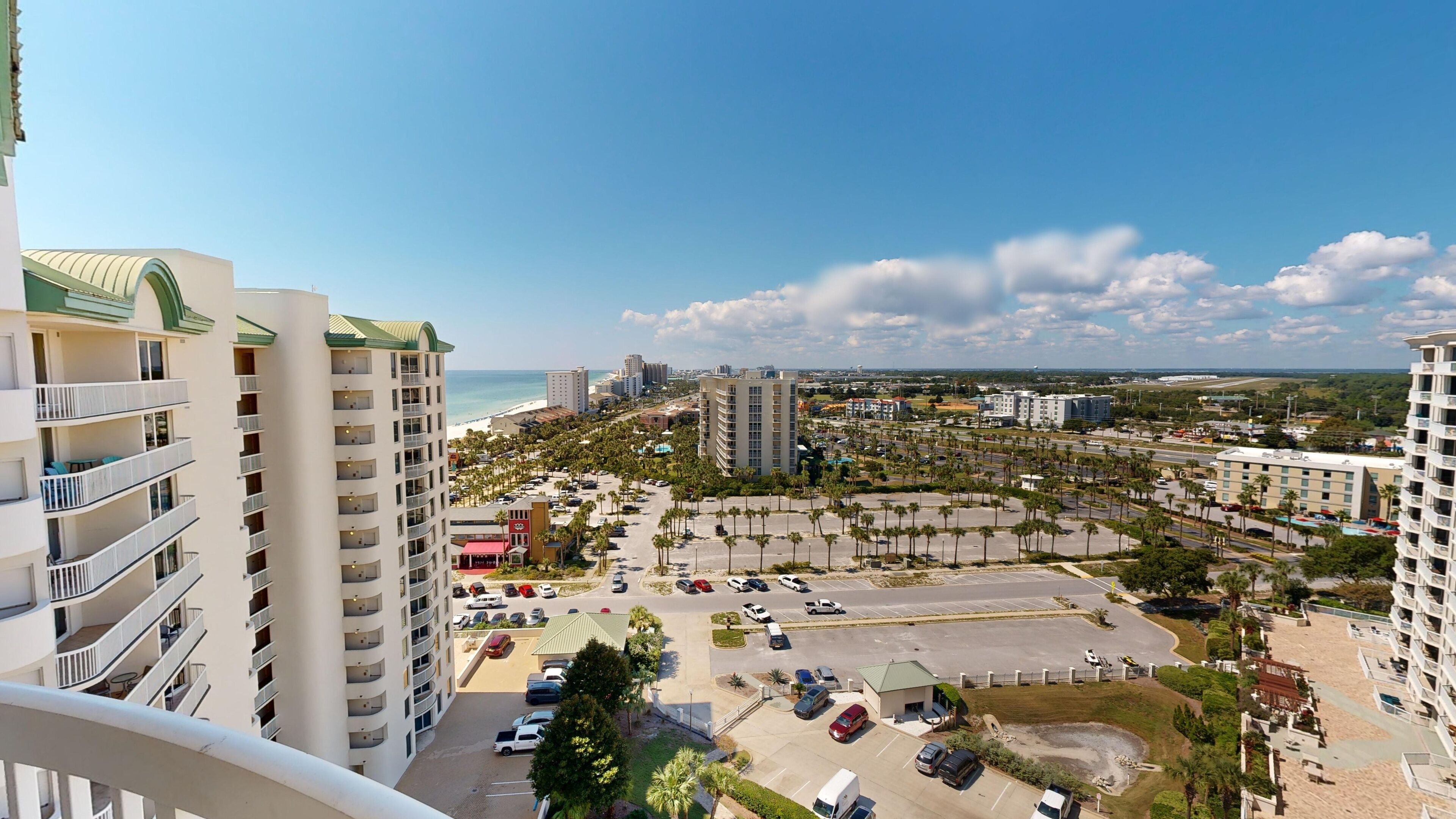 A high-rise hotel with multiple balconies, a parking lot, and a view of the beach and ocean.