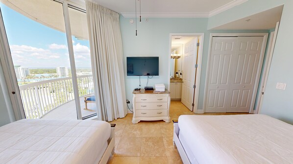 A hotel room with two beds, a TV, a dresser, and a balcony with a view of buildings and greenery.