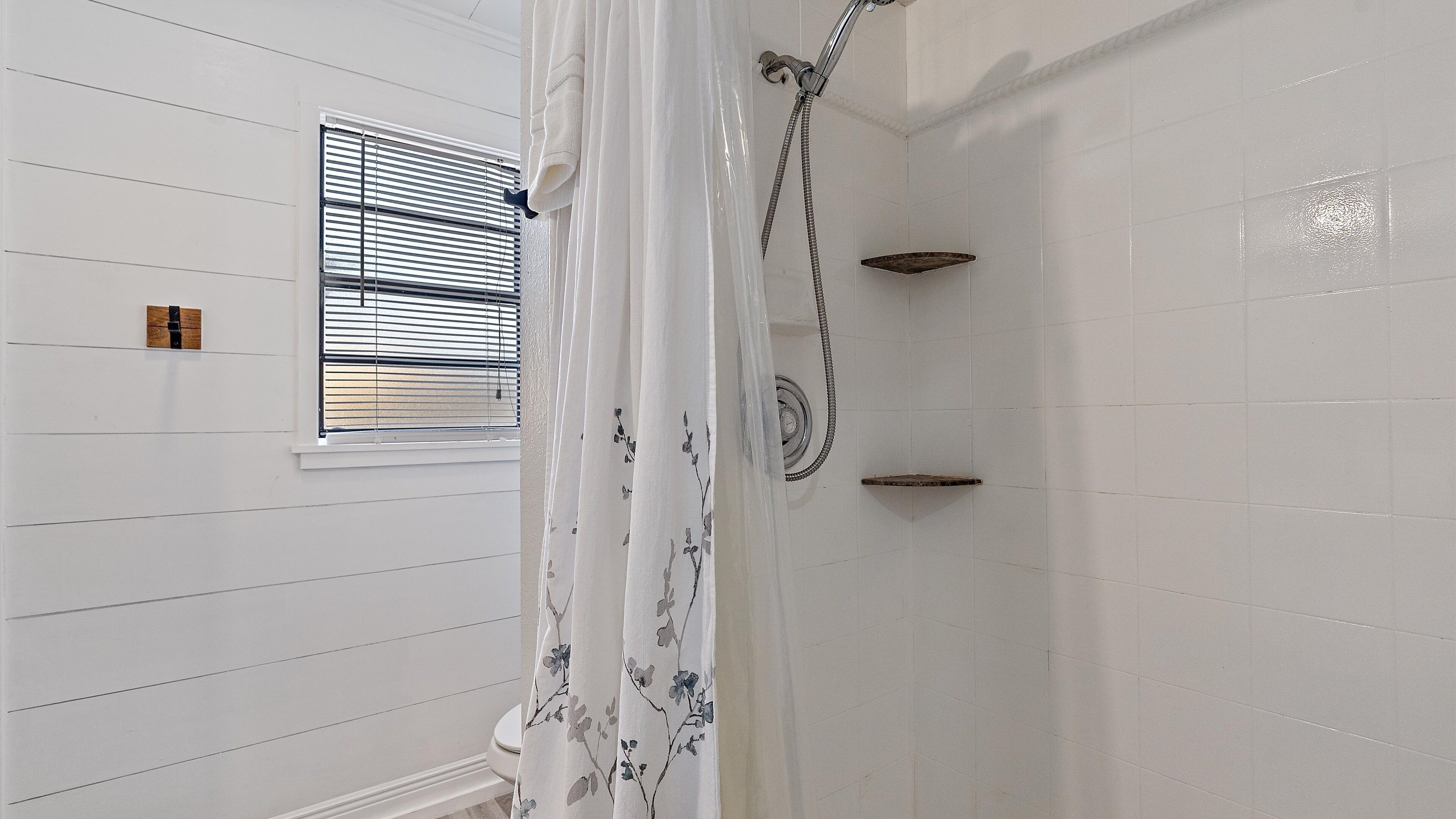Ein Badezimmer mit weißen Wänden, einer Dusche inklusive Handbrause, einem Fenster mit Jalousien und einer kleinen Ablagefläche.