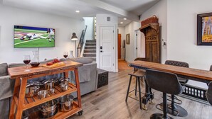 Una cocina moderna con una mesa de madera, taburetes de bar y un televisor mostrando un partido deportivo.
