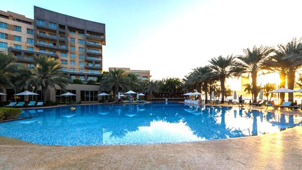 Ein Hotelpoolbereich mit klarem, blauem Pool, Liegestühlen, Sonnenschirmen und Palmen.