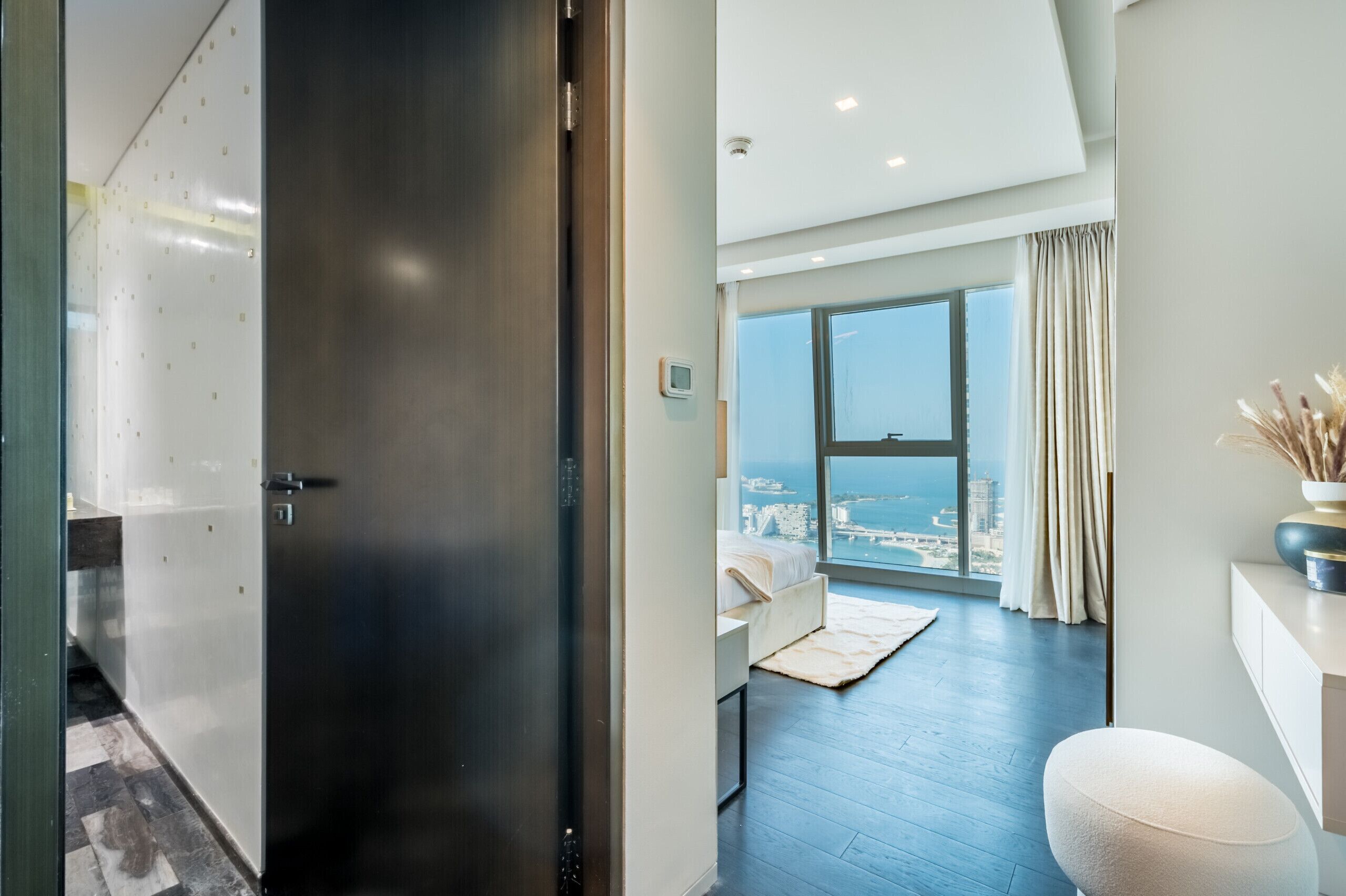 A modern hotel room with a large window, a city view, a dark wooden door, and a white wall.