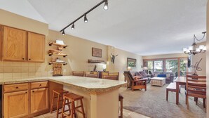 A spacious kitchen with wooden cabinets, a granite countertop, and bar stools. There is a dining area with a table and chairs, and a living room with a sofa and a fireplace. - Beautiful 2 bdrm condo in Breckenridge, Steps to gondola/Main Street! (Breckenridge)