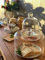 Una mesa de madera con una campana de cristal sobre un plato de pan en rebanadas, un tarro de cristal y un pequeño recipiente con guarnición.