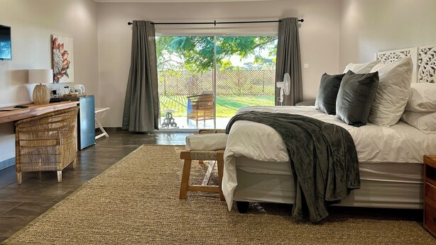 A bedroom with a large bed, a desk, and a chair. There is a view of a garden through the window. - Sun Valley Farm Guest House (Chegutu)